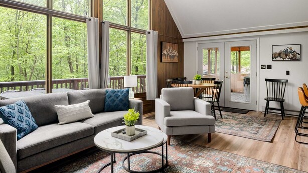 A spacious living area with a large window, a sofa, a chair, a coffee table, and a dining table with chairs. - Cozy chalet w/ forest views & hot tub (Hedgesville)