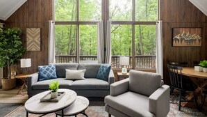 A cozy living room with a gray sofa, a round coffee table, and a gray armchair. Large windows offer a view of a forested area. - Cozy chalet w/ forest views & hot tub (Hedgesville)