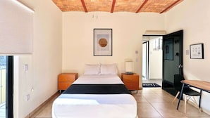 Une chambre d’hôtel comprenant un lit, des tables de chevet, un bureau, une chaise, un ventilateur de plafond, un tableau au mur et une porte donnant sur une autre pièce.