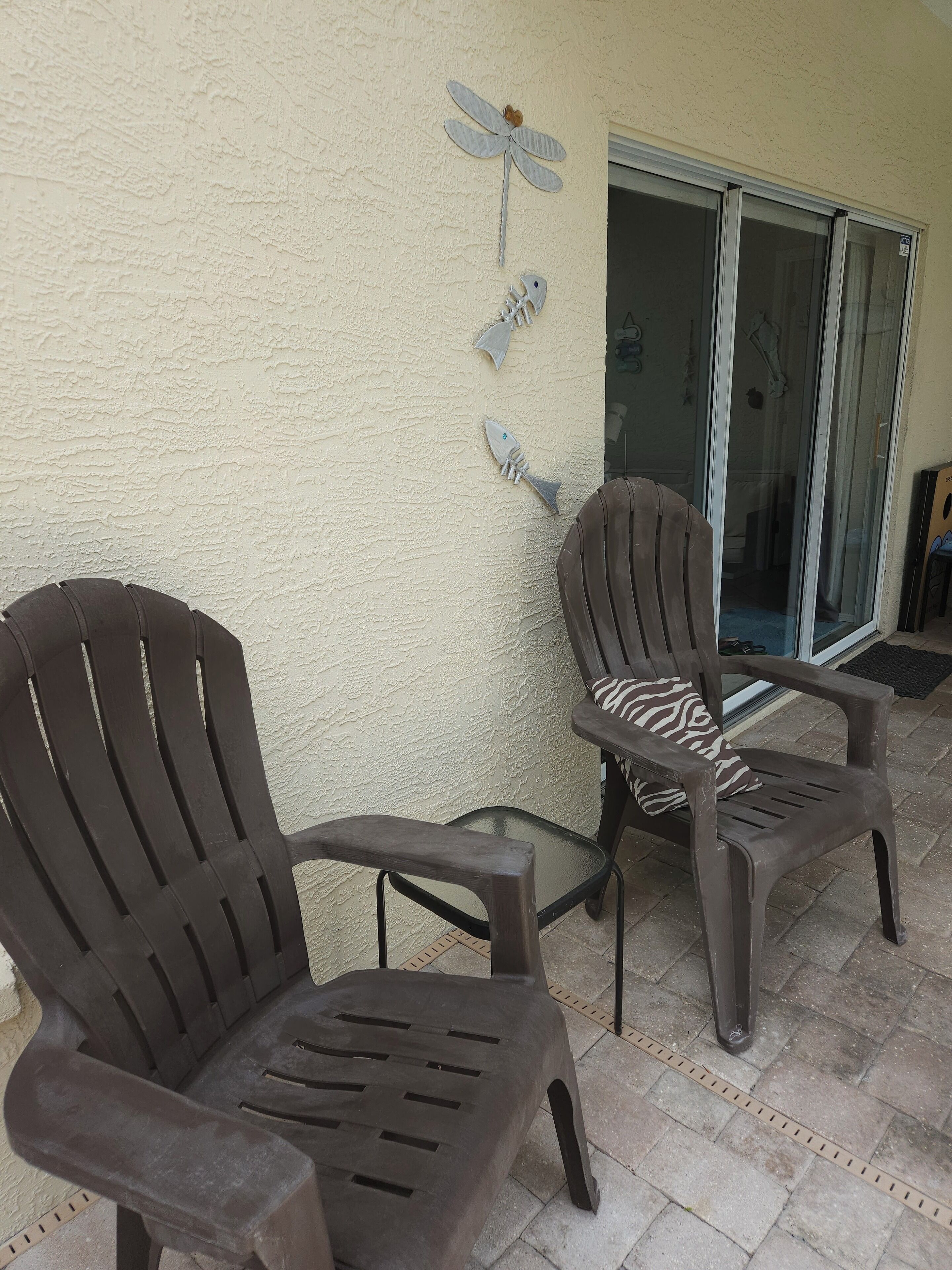 Two plastic chairs with armrests and a glass table on a patio with a textured wall and decorative fish and dragonfly art.