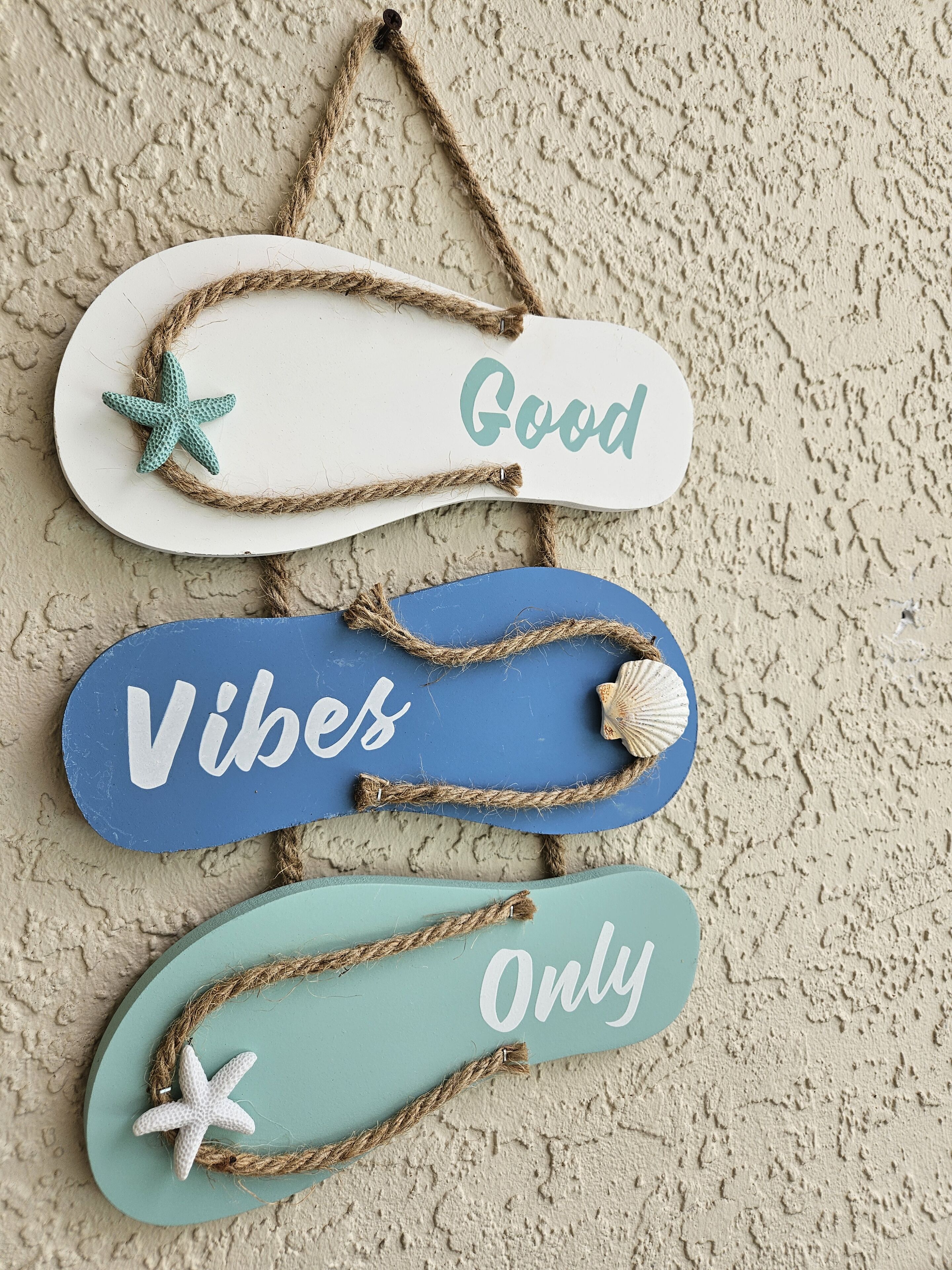 En vægdekoration med tre træskilt, der siger "Good", "Vibes" og "Only", hver med en søstjerne og en søshell.