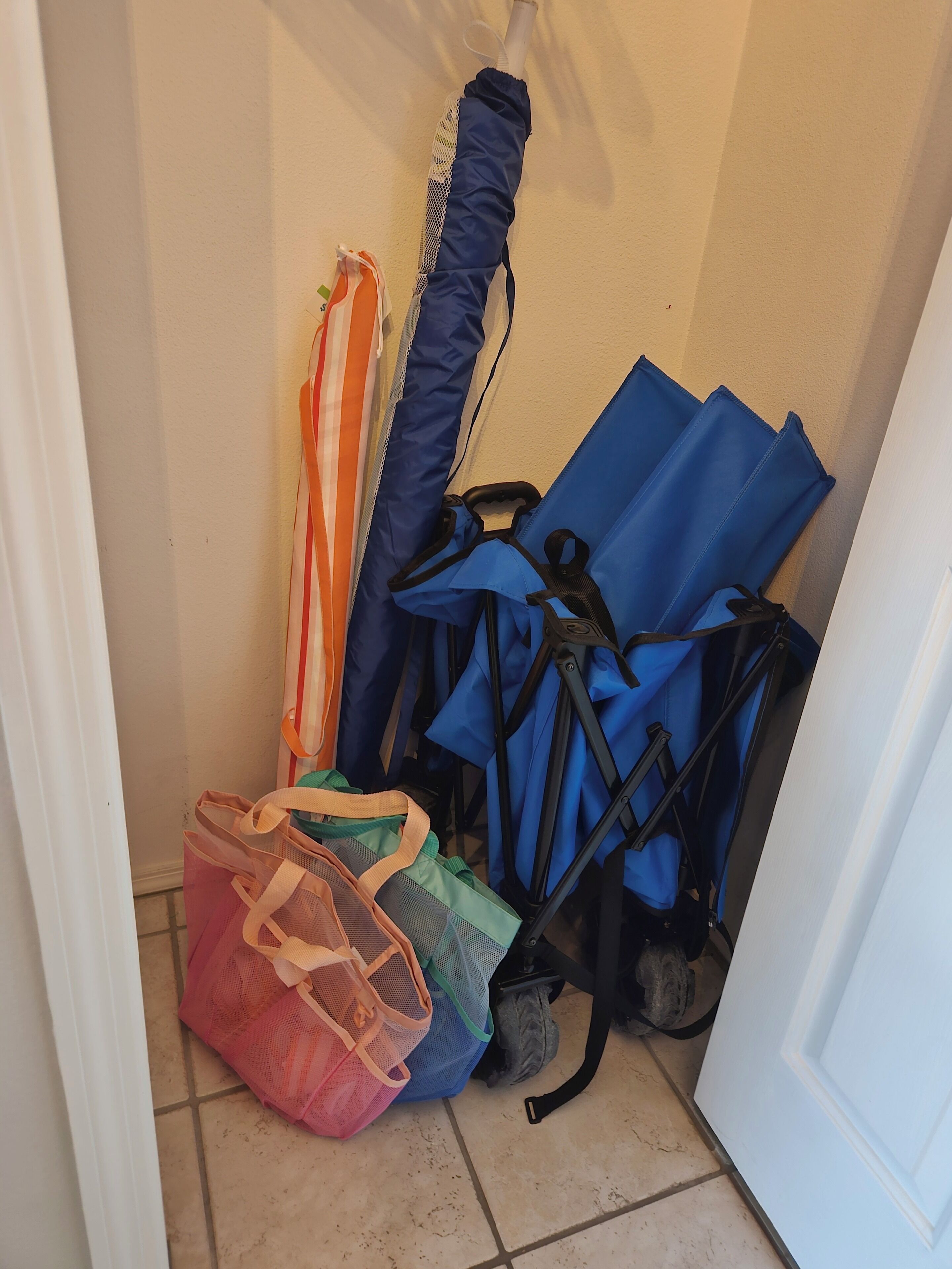 A corner of a room with a tiled floor, a white door, and a wall. There are several bags and umbrellas stacked against the wall.