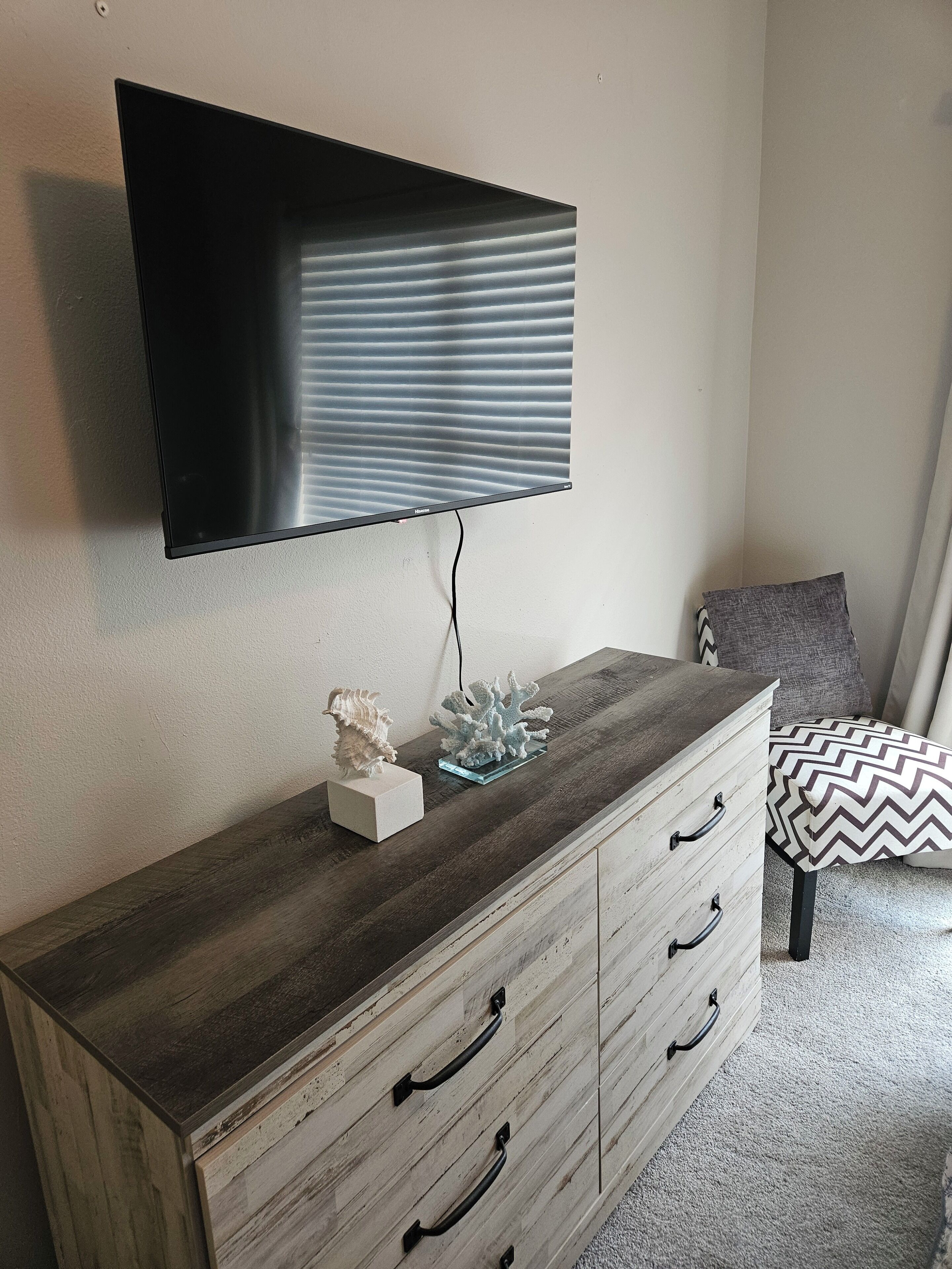 Una habitación con un televisor de pantalla plana montado en la pared, un aparador de madera con cajones, una concha decorativa y un coral sobre el aparador.