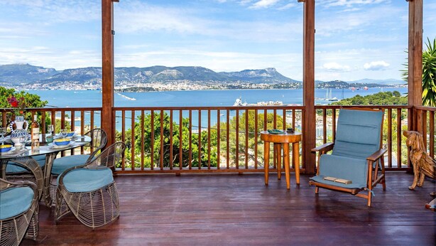Una terraza de madera con sillas de mimbre y una mesa lista para comer, con vistas a una ciudad costera y montañas al fondo.