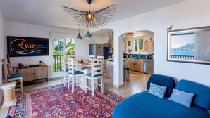 Una sala de estar moderna con un sofá azul, un comedor con alfombra estampada, y una cocina visible a través de una puerta abierta.