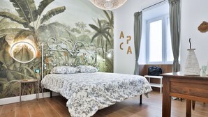 Un dormitorio con cama, un mural en la pared, una mesita de noche, una lámpara y una ventana con cortinas.