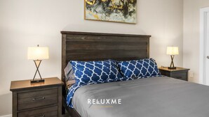 A bedroom with a dark wooden headboard, two bedside tables with lamps, a bed with patterned pillows, and a painting above the bed. - Reluxme | Uptown 3BR w/ King Beds & Spa Access (Charlotte)