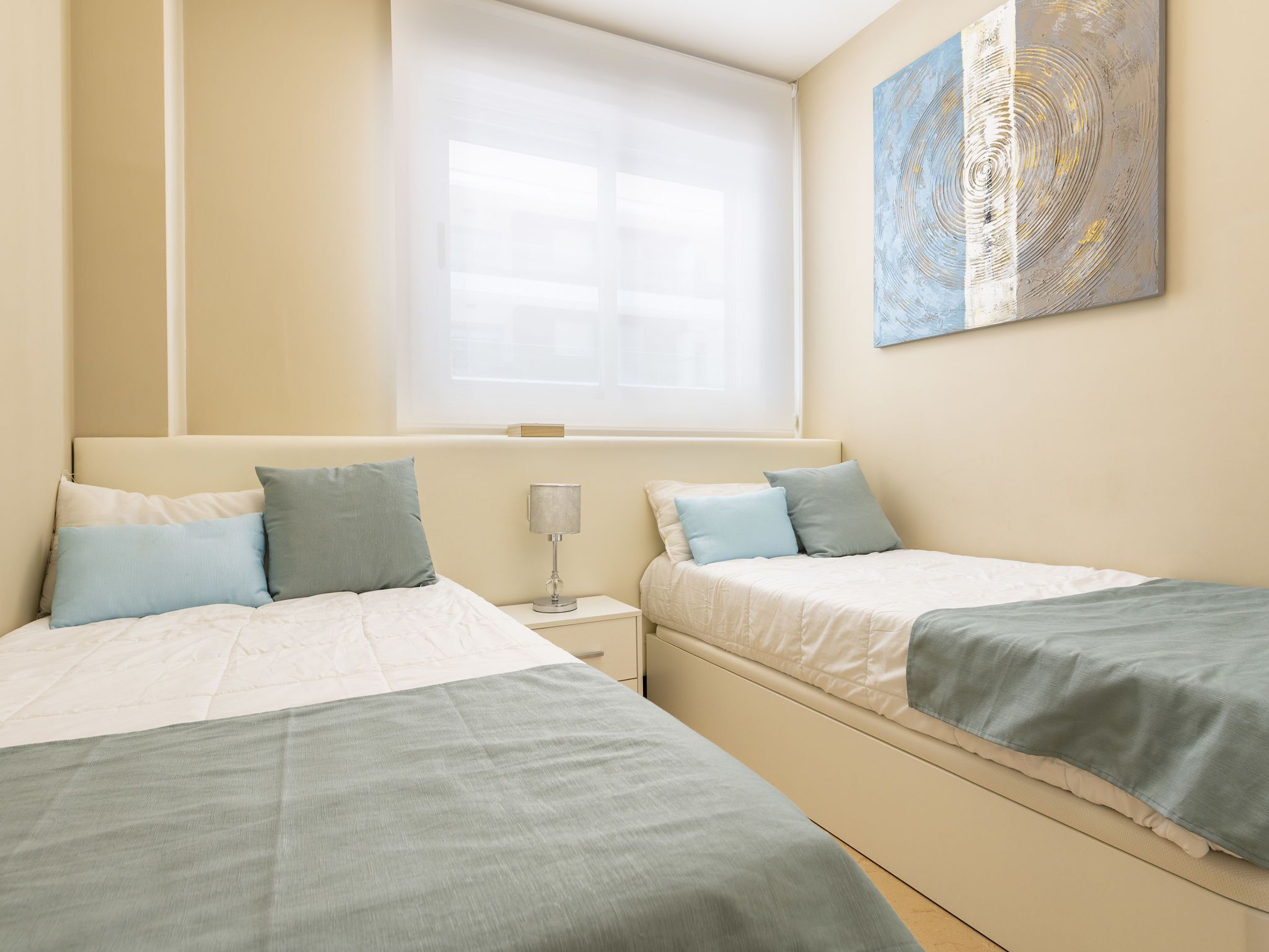 A hotel room with two beds, a window with blinds, a bedside table with a lamp, and a piece of abstract art on the wall.
