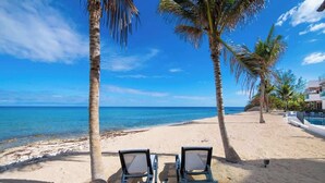 A beach with two lounge chairs facing the ocean, palm trees, and a clear blue sky.