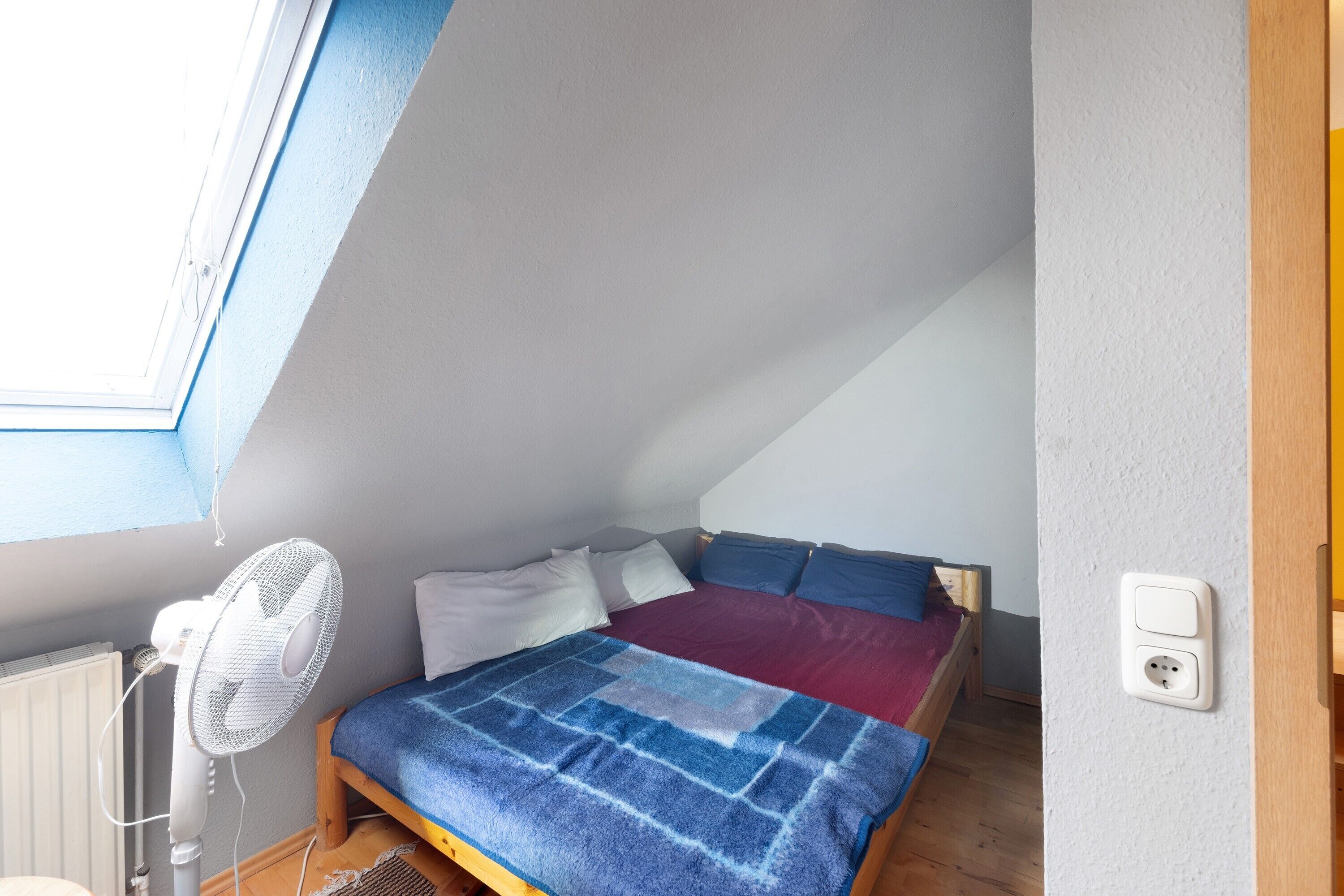 A bedroom with a sloped ceiling, a bed with a blue and purple blanket, a standing fan, and a window.