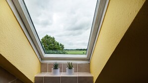 Um claraboia com duas plantas em vasos, vista para uma paisagem verde e uma parede amarela.