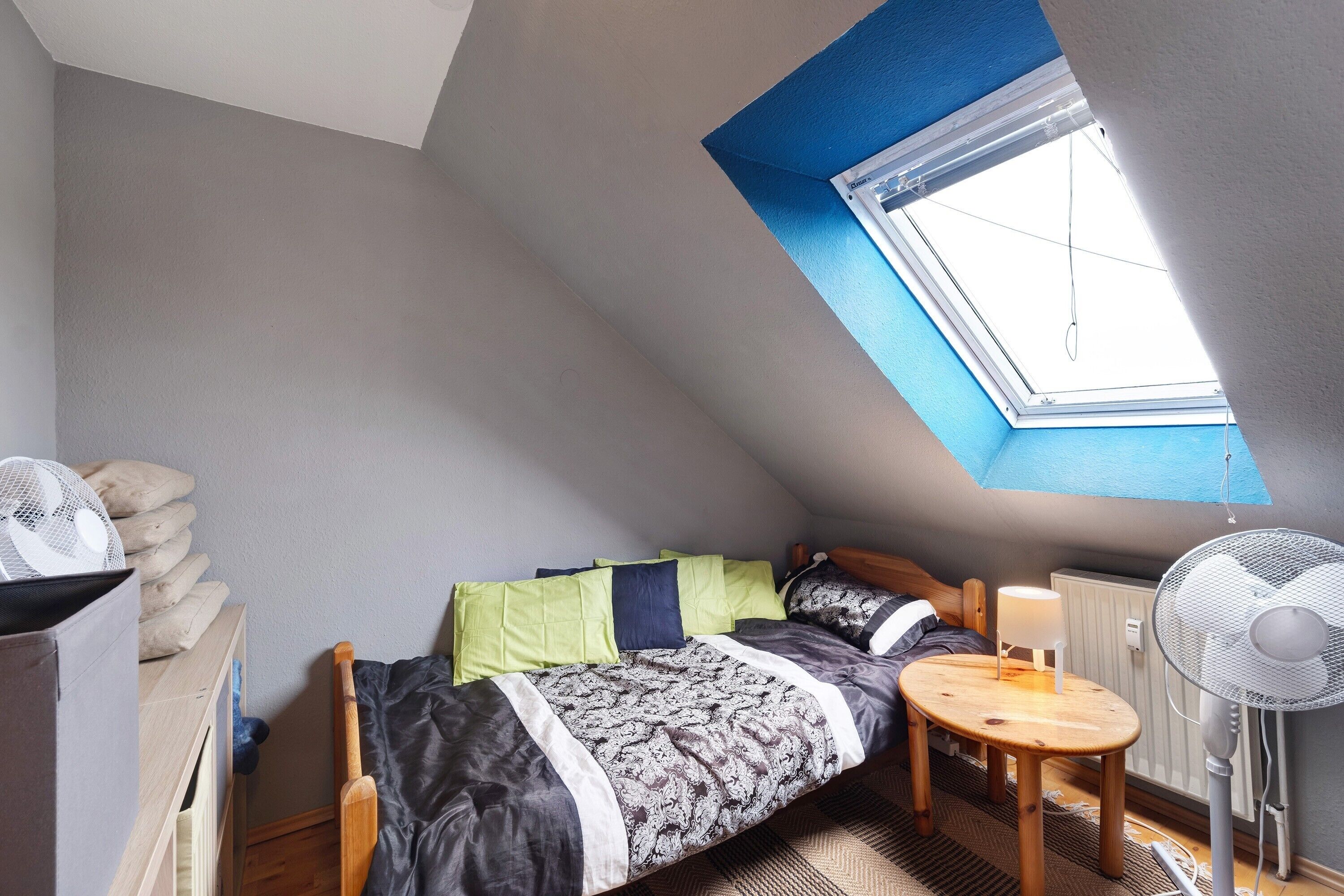 Ein kleiner, gemütlicher Raum mit schrägem Dach, einem Oberlicht, einem Bett mit gemustertem Überwurf, einem Nachttisch mit Lampe und einem Standventilator.