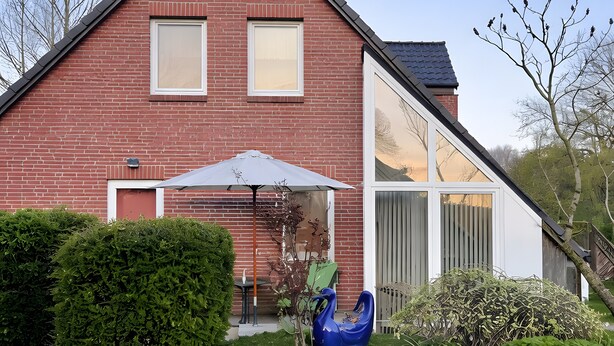 Ein zweistöckiges Backsteinhaus mit geneigtem Dach, einer überdachten Terrasse mit Tisch und Stühlen sowie einem kleinen Garten mit gelben Blumen und grünen Sträuchern.