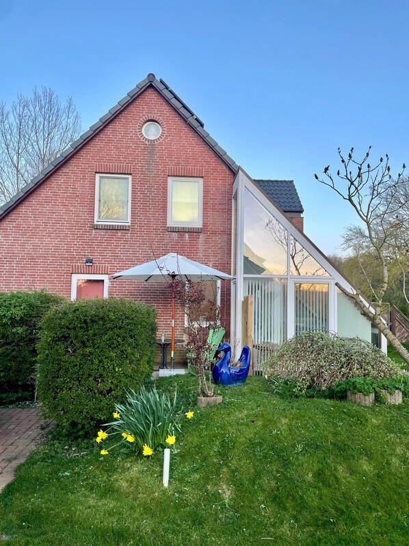 A red brick house with a white balcony, a blue sculpture, and a garden with yellow flowers.