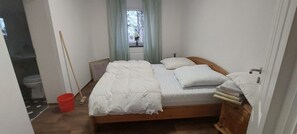 A bedroom with a bed, a window with curtains, a wooden floor, and a red bucket.