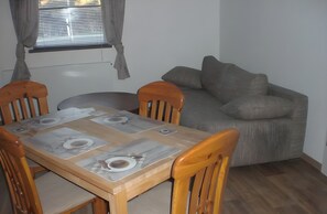 A dining table set with placemats, plates, and cutlery, flanked by wooden chairs, with a grey sofa and a window with blinds in the background.