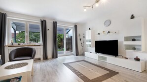 A modern living room with a TV, white furniture, a large window, and a view of greenery outside.