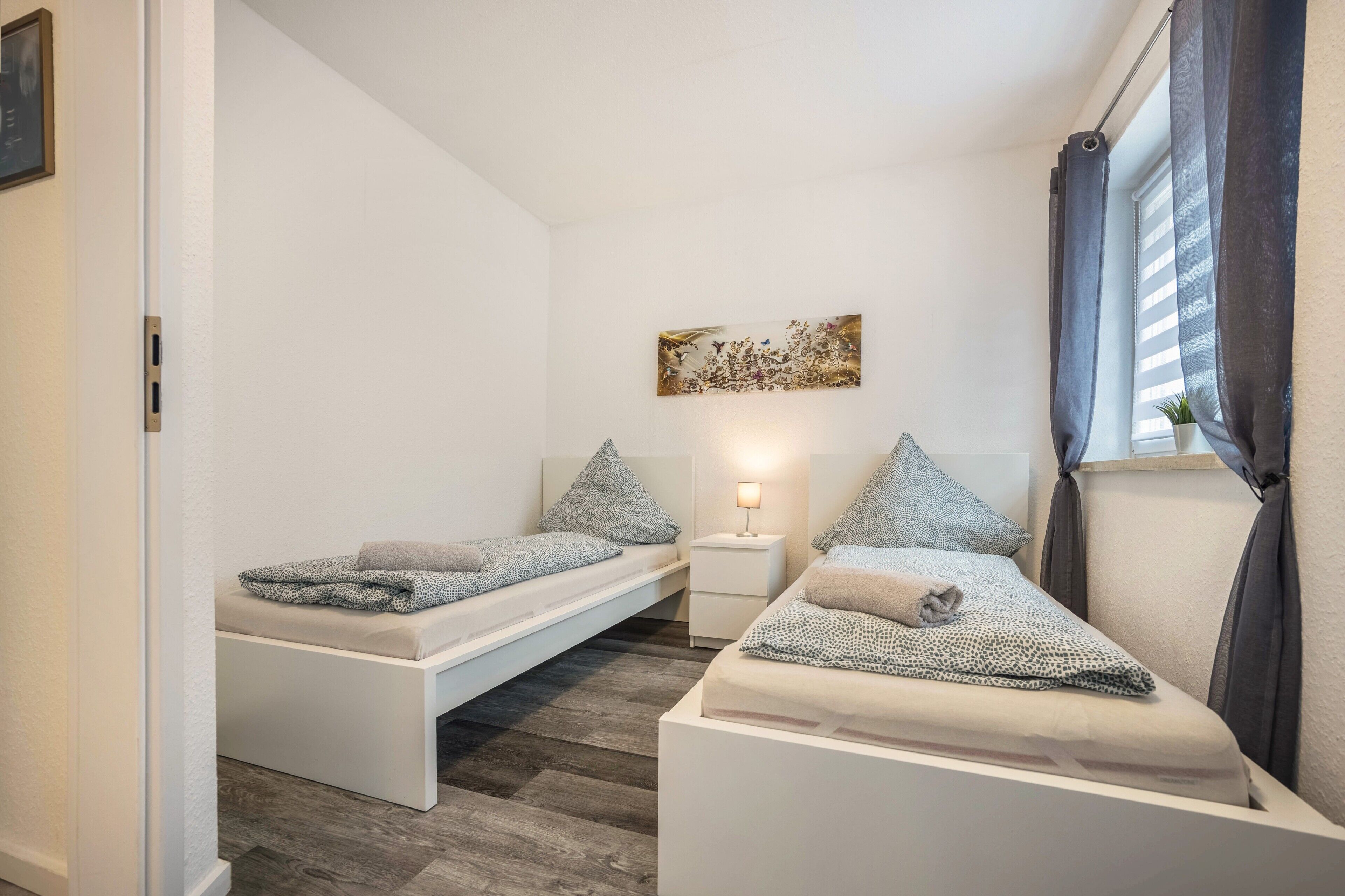 Un dormitorio pequeño y moderno con dos camas individuales, una mesita de noche con lámpara y un cuadro en la pared.