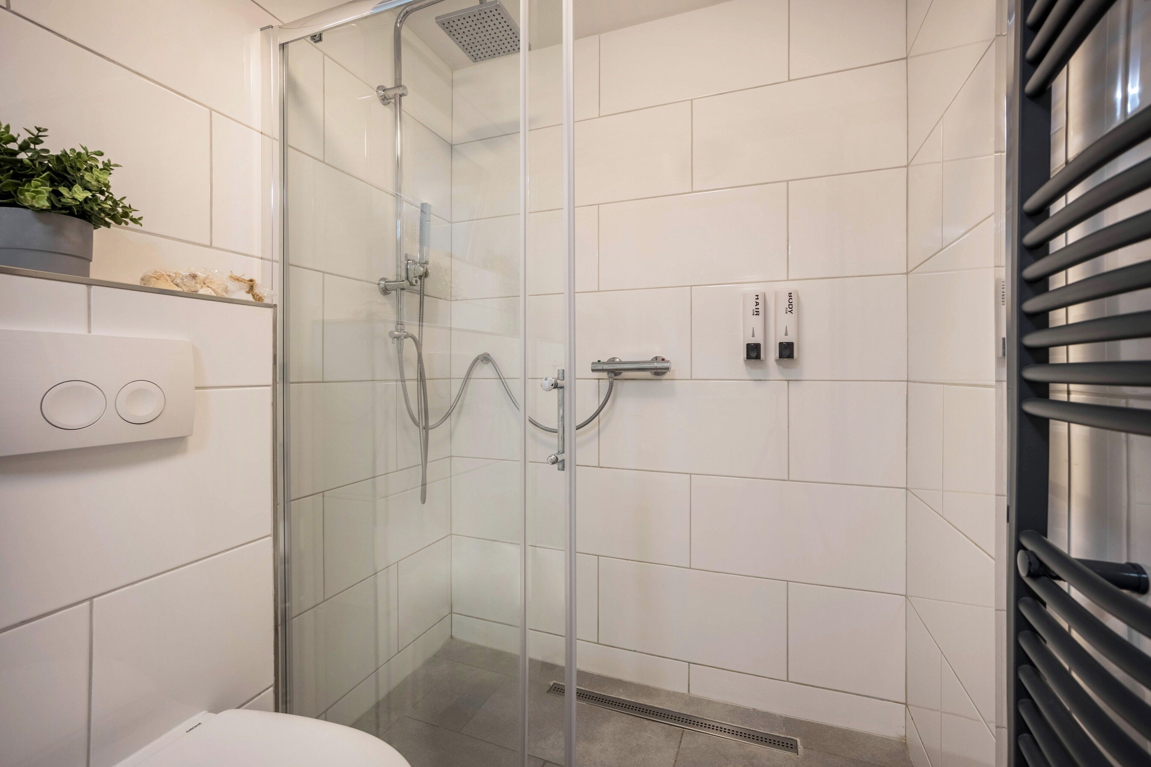 Uma casa de banho moderna com cabine de duche em vidro, azulejos brancos, sanita, lavatório com torneira e uma pequena estante com uma planta.