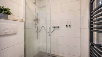 Uma casa de banho moderna com cabine de duche em vidro, azulejos brancos, sanita, lavatório com torneira e uma pequena estante com uma planta.