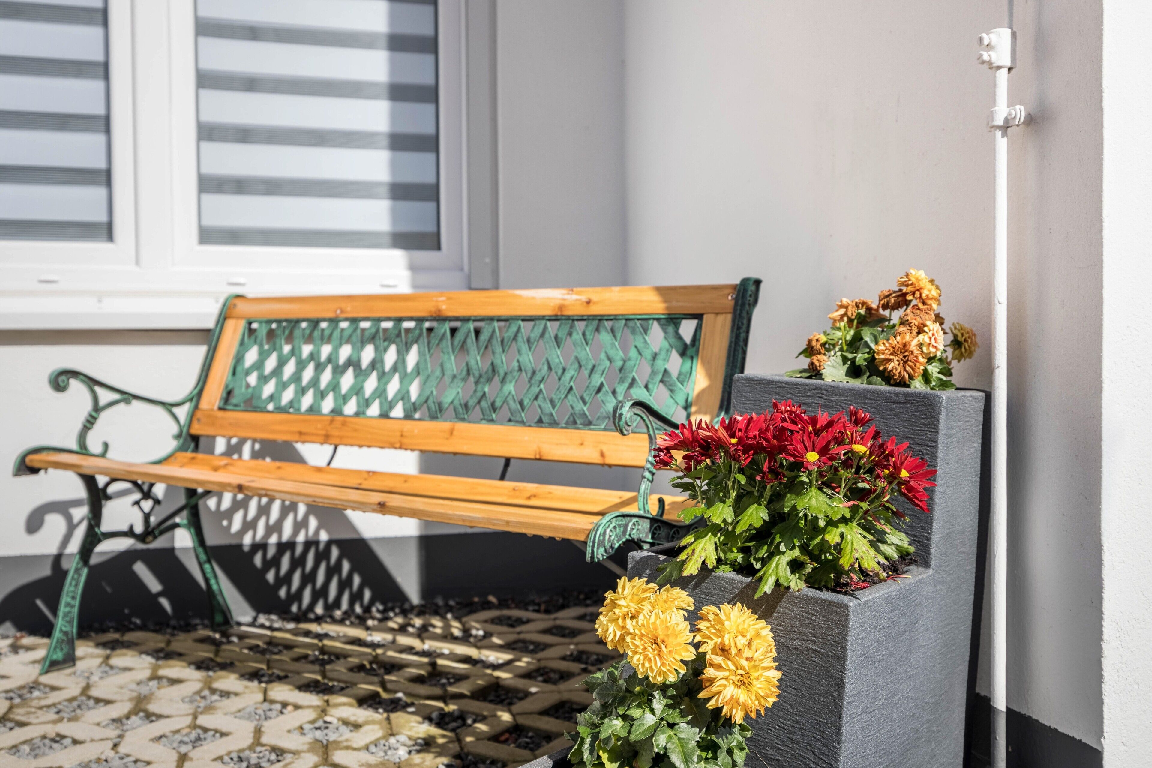 A bench with a green metal frame and wooden slats, positioned next to a wall with a potted plant containing yellow and red flowers.