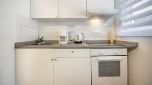 Une cuisine moderne avec des meubles blancs, un four encastré, une machine à café et une fenêtre équipée de stores.