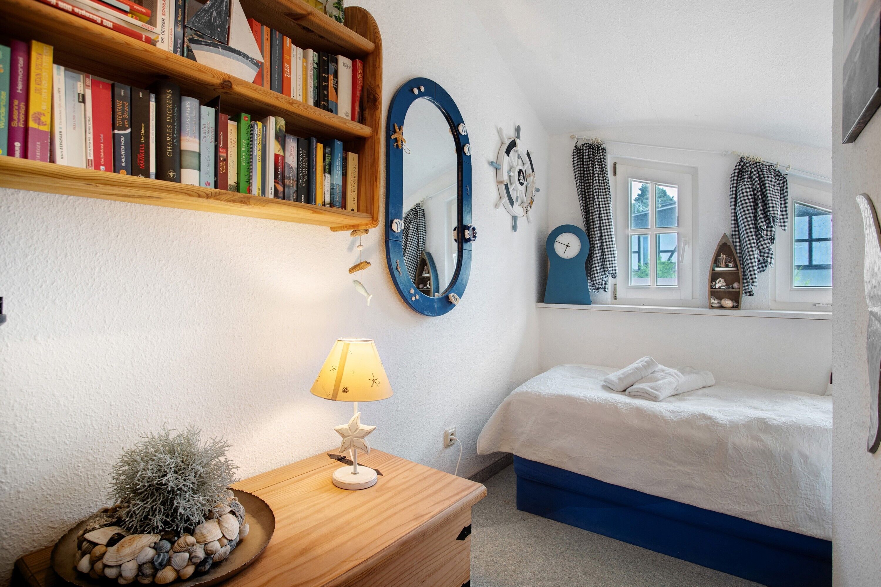 A bedroom with a bed, a bedside table with a lamp, a mirror, a bookshelf, and a window with curtains.