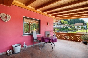 Une terrasse couverte avec une table et des chaises, une fenêtre donnant sur des maisons et un décor en forme de cœur sur le mur.