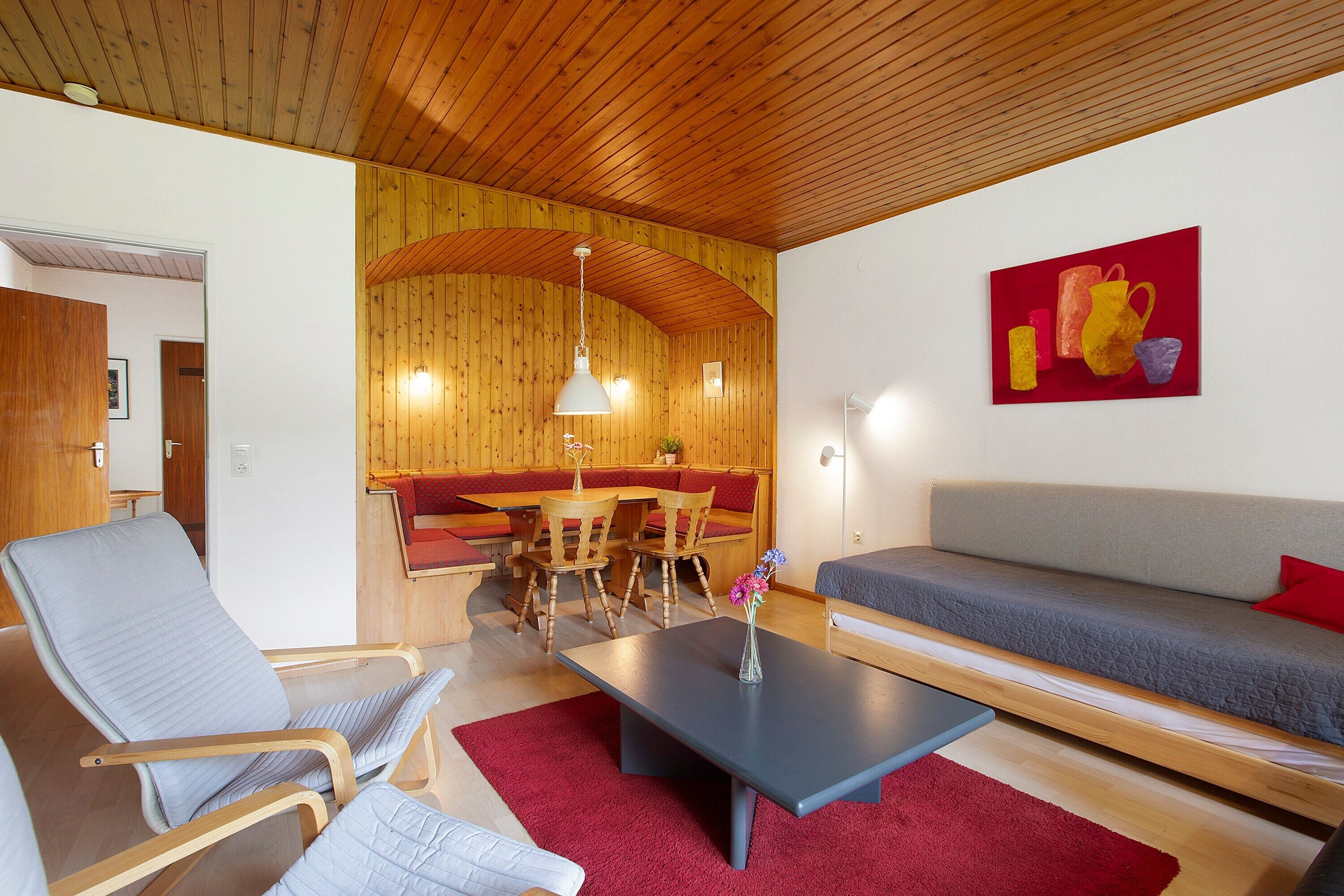 A room with a sofa, a chair, a table, a red rug, wooden walls, a wooden ceiling, and a painting on the wall.