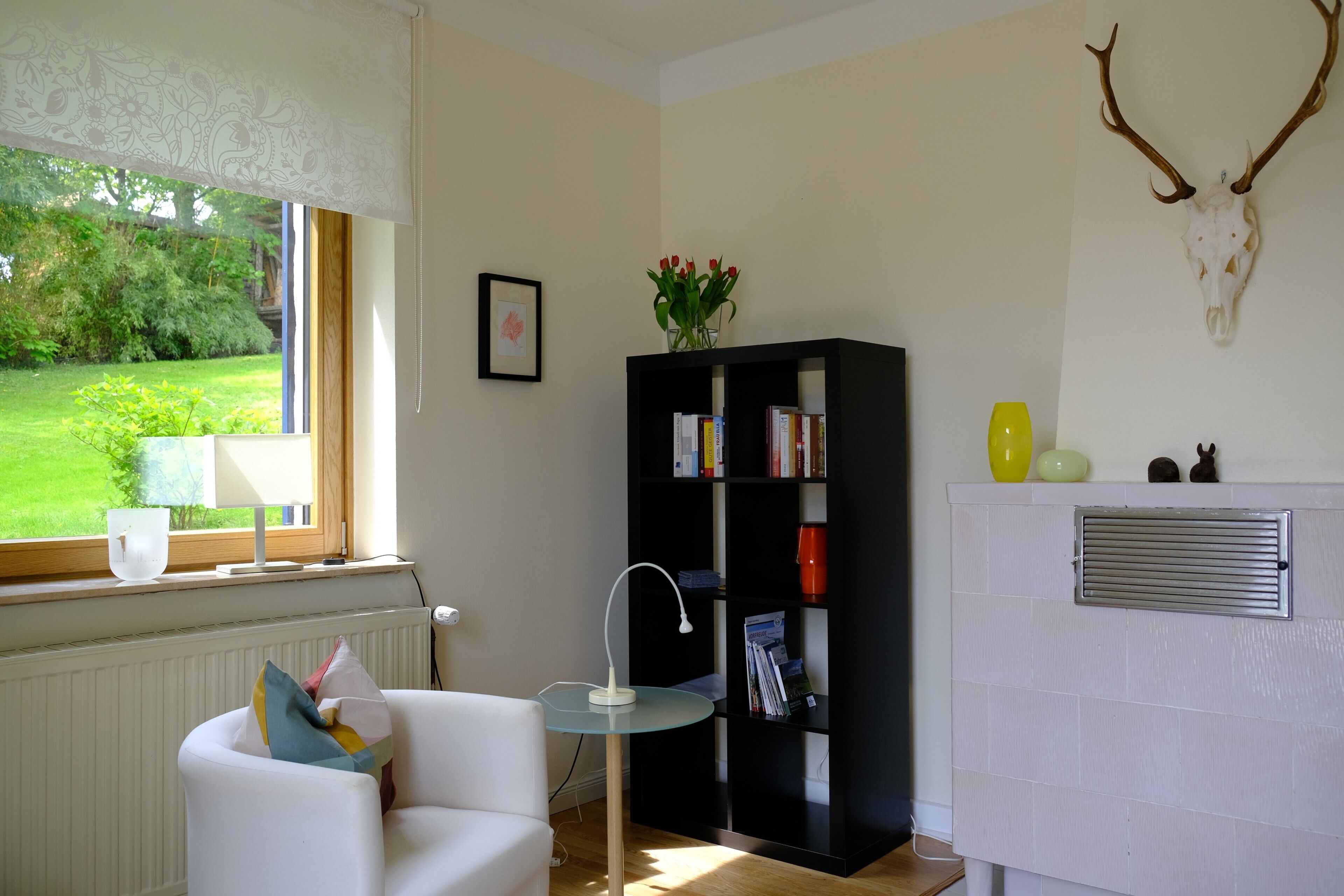 Et værelse med en hvid radiator, en sort reol, en hvid stol med farverige puder, et lille bord med en lampe, og et vindue med udsigt over grøn vegetation.