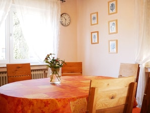 Une salle à manger avec une table ronde, des chaises, une horloge, des œuvres d’art encadrées et une fenêtre avec des rideaux.