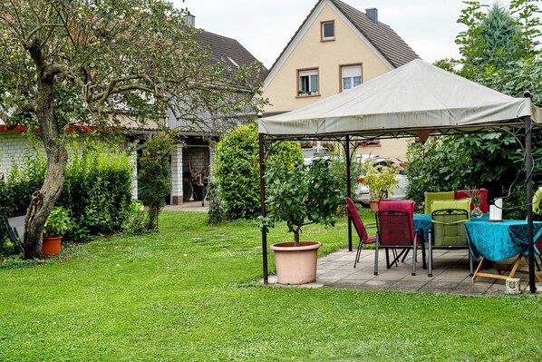 Un jardin avec une table et des chaises sous une tente, entouré de verdure et d’une maison.