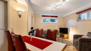 Un comedor con una mesa, sillas y un mantel individual rojo. Se ve un sofá, un televisor y una ventana con persianas en el fondo.