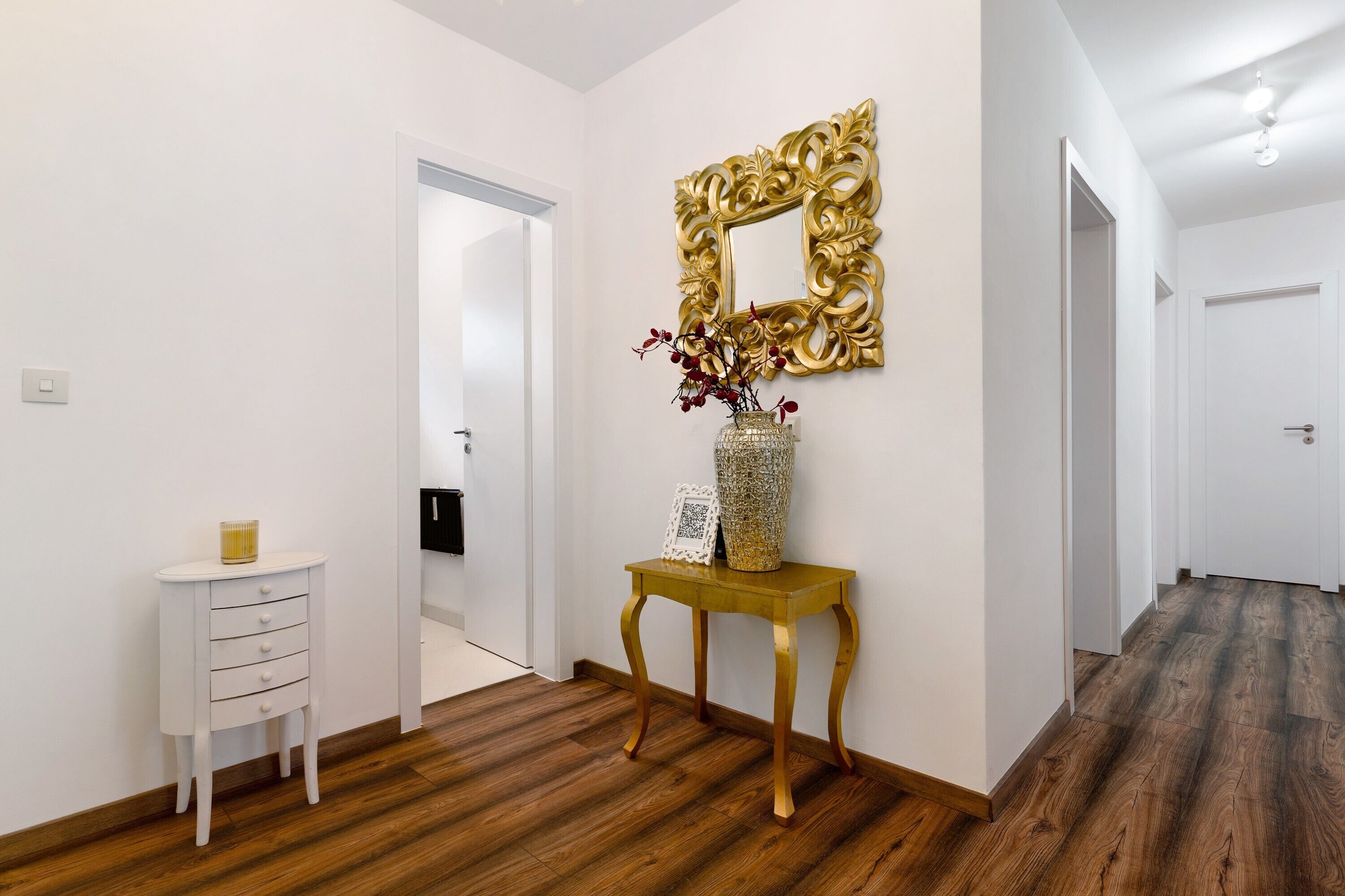 Un couloir avec un mur blanc, un plancher en bois, une table d’appoint blanche avec des tiroirs, un miroir doré et un vase rempli de fleurs.