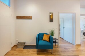 Una habitación con sofá azul, estante de madera, luz empotrada en la pared, planta y zona de lavandería.
