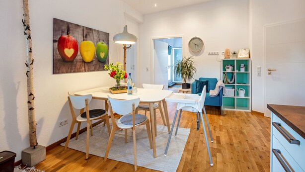 A modern kitchen with white cabinets, a dining table with chairs, a hanging light fixture, and a painting of apples on the wall.