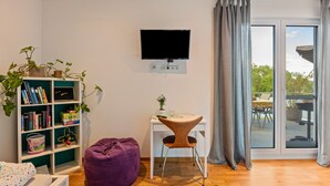 A modern living room with a flat-screen TV, a white desk, a purple bean bag, a bookshelf, and a sliding glass door leading to an outdoor area.