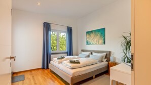 A bedroom with a bed, a bedside table, a window with curtains, a painting on the wall, and a plant.