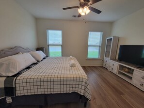 Un dormitorio con cama, televisor, ventilador de techo y dos ventanas.