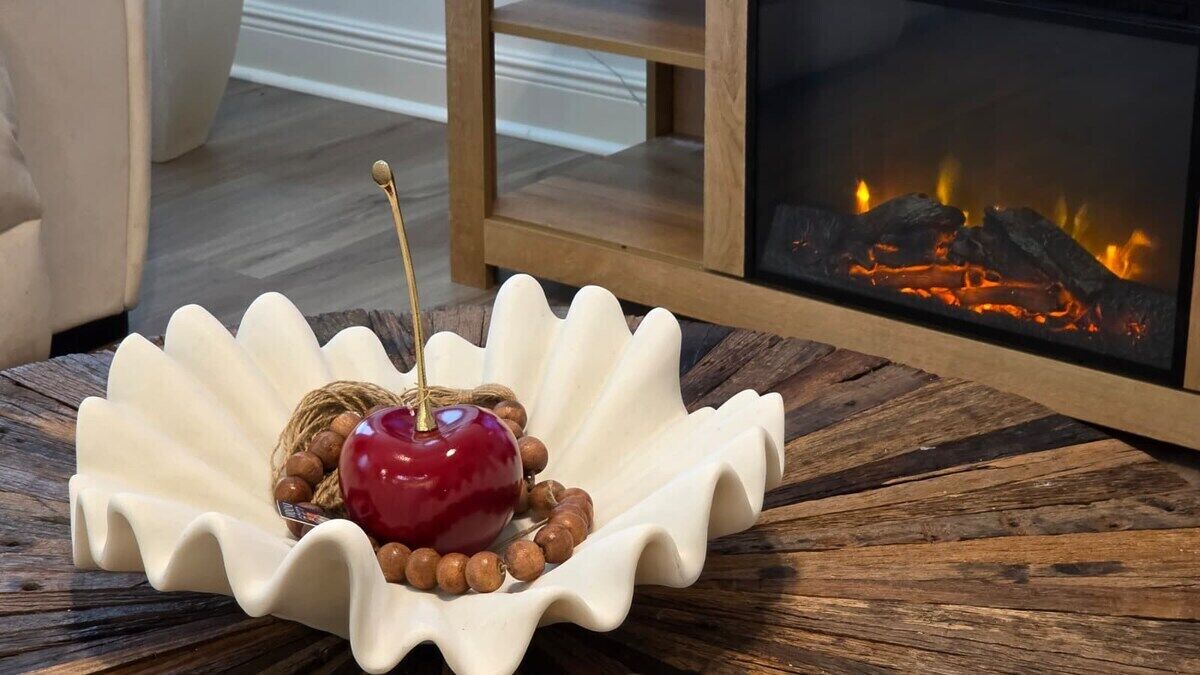 Un plateau décoratif en forme de coquillage blanc, sur lequel reposent une pomme rouge et des noix, est posé sur une table en bois. On aperçoit une cheminée en arrière-plan.