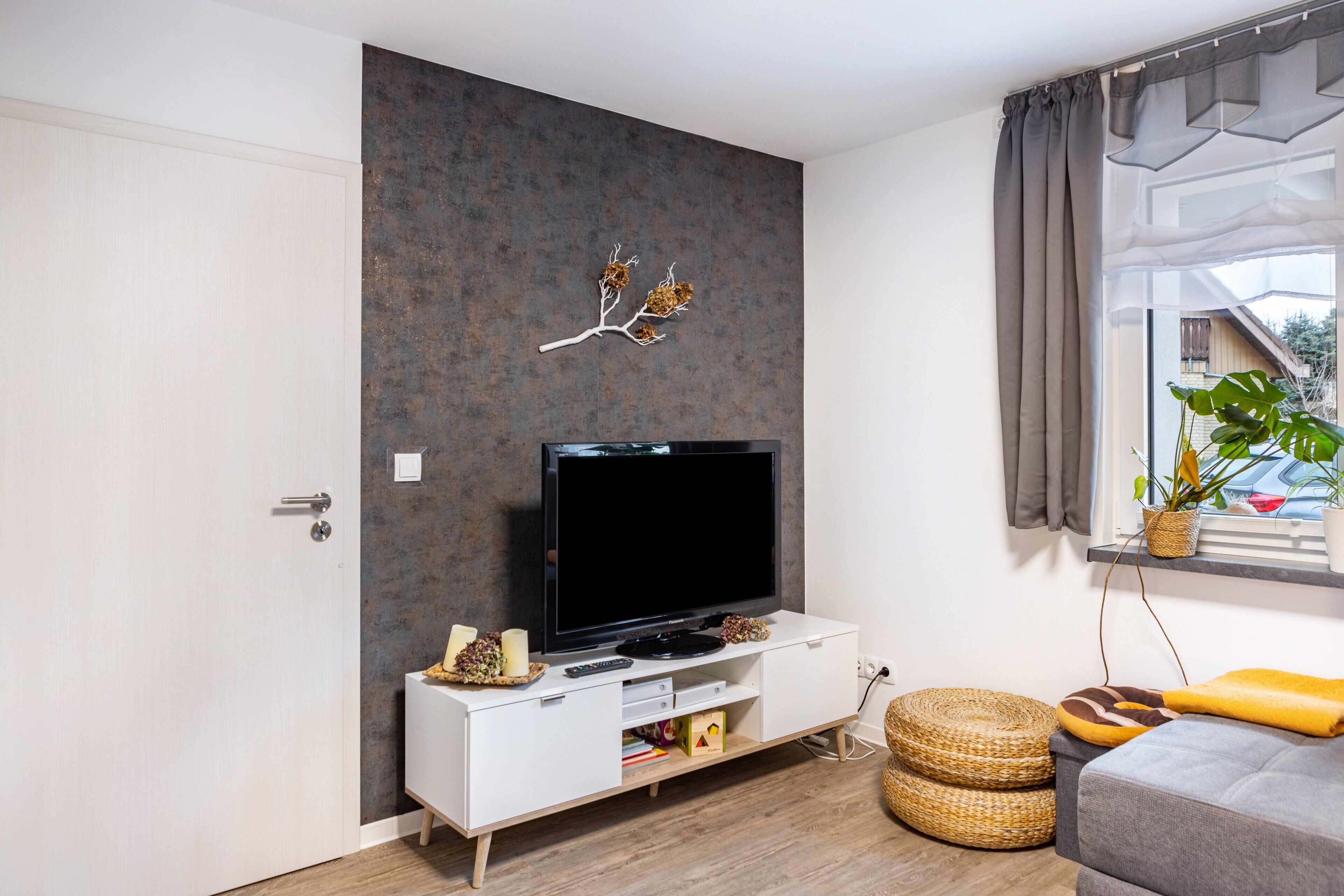 A modern living room with a flat-screen TV on a white console, a decorative wall piece, a window with curtains, and a plant.