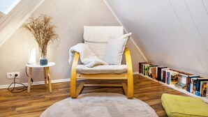 Une pièce mansardée chaleureuse, avec un fauteuil à bascule en bois, une petite table d’appoint surmontée d’un vase, une bibliothèque et un tapis moelleux.