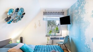 Une chambre à coucher avec un lit, un bureau, une chaise, une télévision et une fenêtre ornée de plantes.