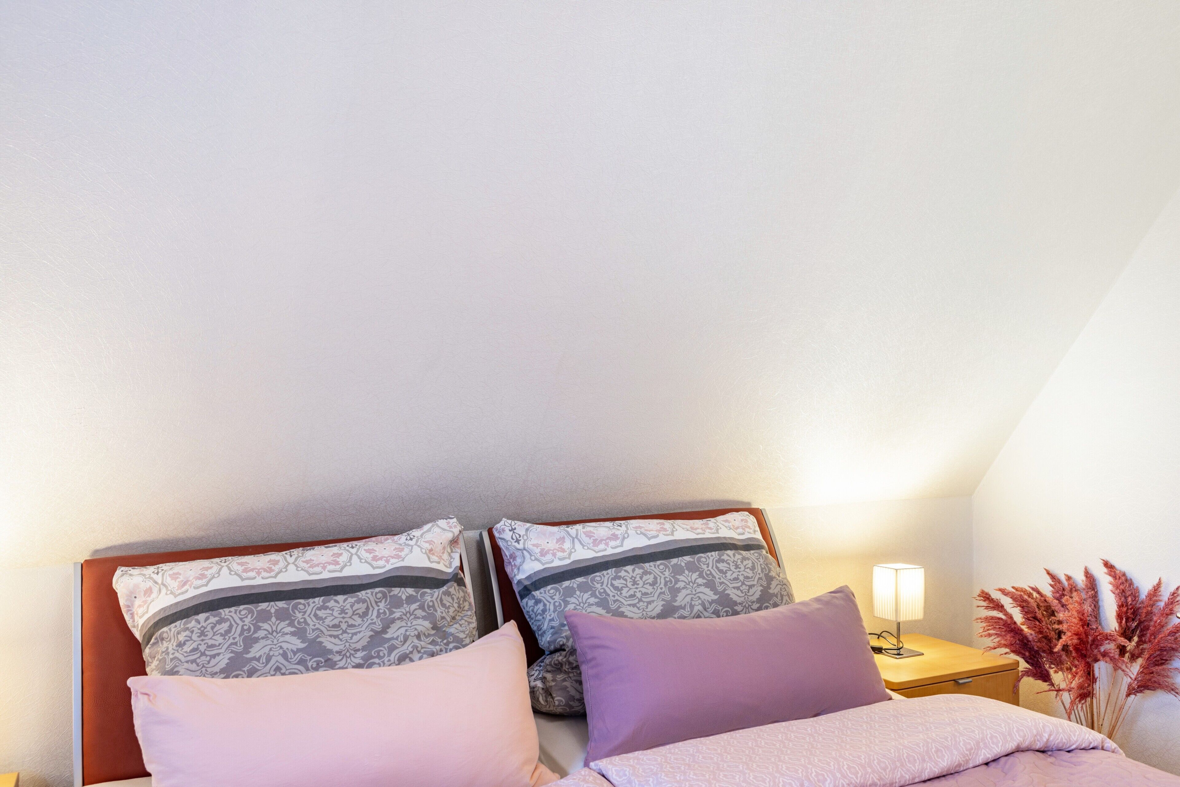 Un dormitorio con cama, almohadas, una mesita de noche con lámpara y un jarrón con flores secas.