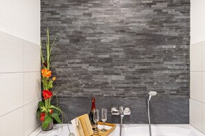 A bathroom with a stone wall, a flower arrangement, a tray with a bottle and glasses, and a shower head.