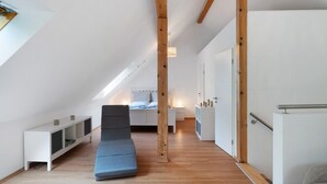 Ein modernes Schlafzimmer im Loft-Stil mit schrägem Dach, Holzbalken, einem grauen Sofa, einem weißen Nachttisch und einer kleinen Regaleinheit.