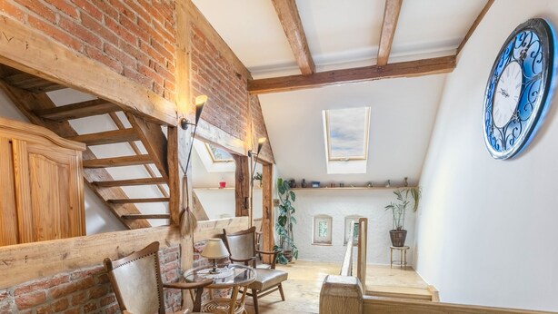 A loft-style room with exposed wooden beams, a brick wall, a wooden staircase, and a large blue clock.