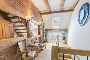 Een loft-achtige kamer met zichtbare houten balken, een bakstenen muur, een houten trap en een grote blauwe klok.