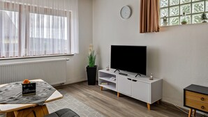 A modern living room with a flat-screen TV, a dining table with a placemat, a potted plant, and a window with curtains.
