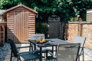 Een terras buiten, met een houten tuinhuis, een metalen hek en een bakstenen muur.